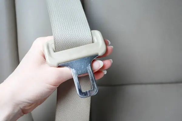 A hand holding a beige seatbelt and its metal tongue beside a vehicle seat
